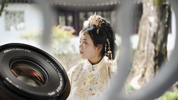 Woman in ornate white and gold traditional dress with elaborate gold headdress, photographed through a camera lens in soft natural light.