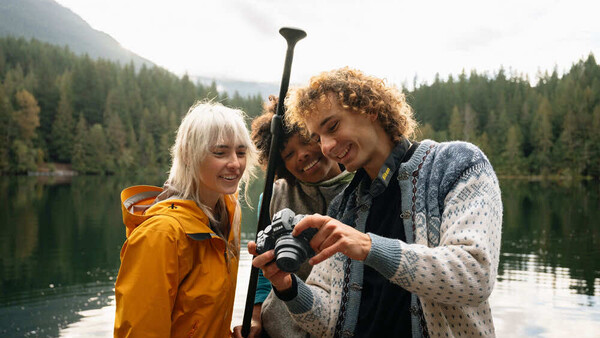 Three people holding a paddle while standing in a boat on a calm alpine lake surrounded by forested mountains