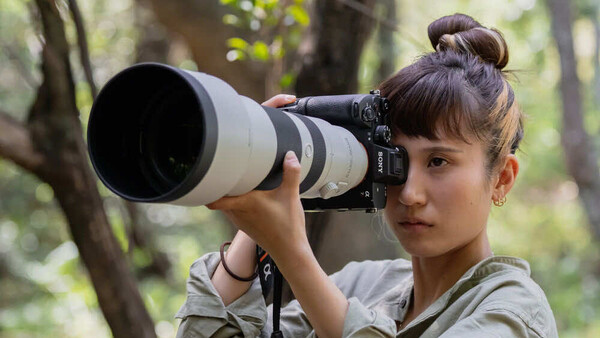 Young woman holding a telephoto lens to her eye in a wooded outdoor setting