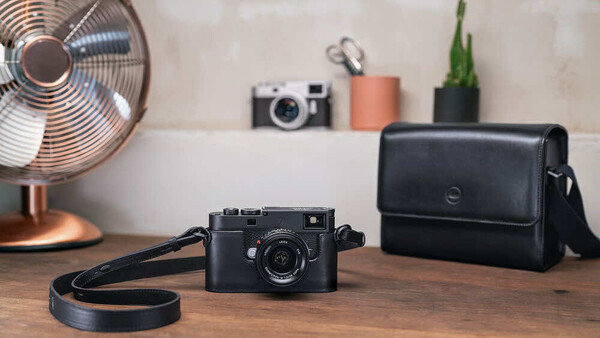 Black rangefinder camera with leather strap on wooden desk beside camera bag
