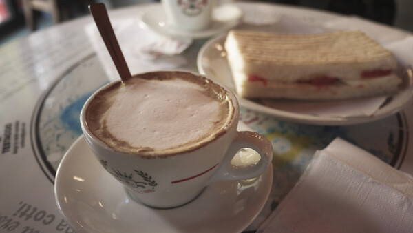 Cappuccino with latte art in a white cup and saucer, with a pastry sandwich on a plate in soft daylight