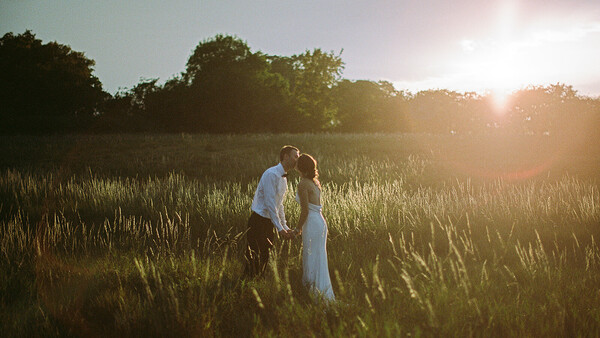 Couple kissing in golden hour sunlight amid tall grass field with trees silhouetted in background