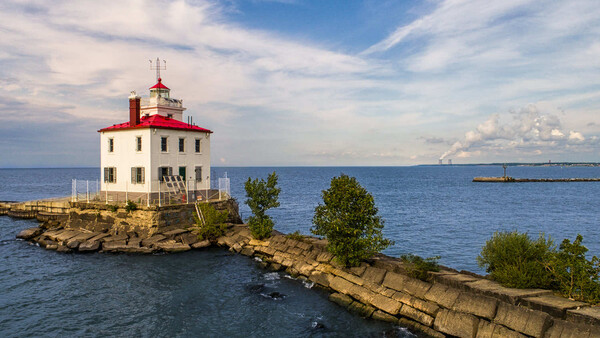 Red and white lighthouse on rocky breakwater surrounded by blue water under partly cloudy sky