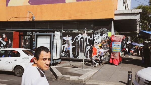Man in white shirt posing on urban street with graffitied storefront and parked vehicles
