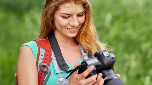 Young woman with reddish-brown hair holding a DSLR camera, wearing a teal top and red backpack outdoors
