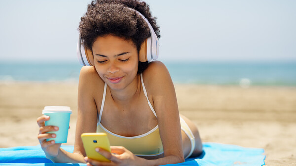 Woman wearing headphones lying on beach towel holding smartphone and beverage