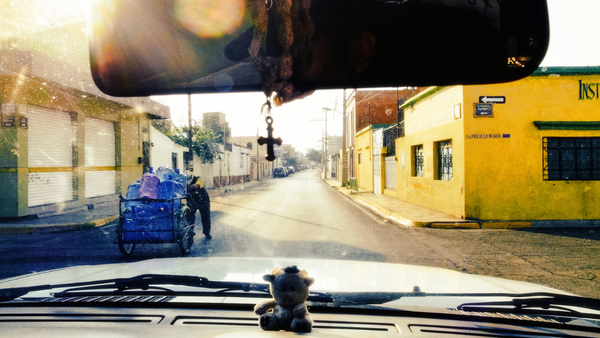 Dashboard view through a vehicle windshield looking down a colorful street with blue cargo tricycle and yellow buildings