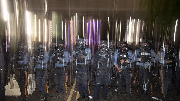 Row of riot police in tactical gear holding shields and batons, illuminated by vertical neon light tubes