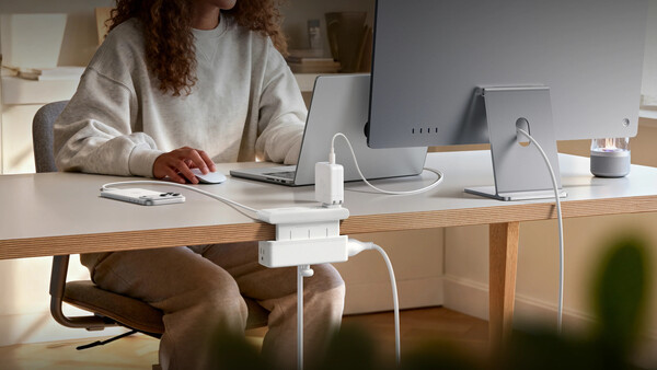 Woman at wooden desk working on laptop surrounded by white tech accessories and charging devices