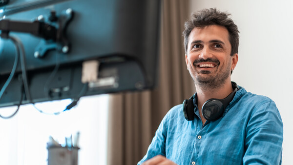 Smiling man in denim shirt at desk with monitor and headphones around neck