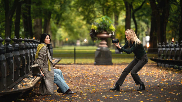 Two women posing playfully on a tree-lined park path with autumn leaves