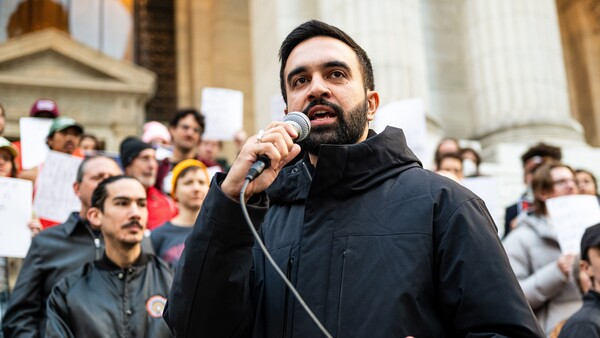 Man with dark beard speaking into microphone at outdoor public gathering with crowd in background