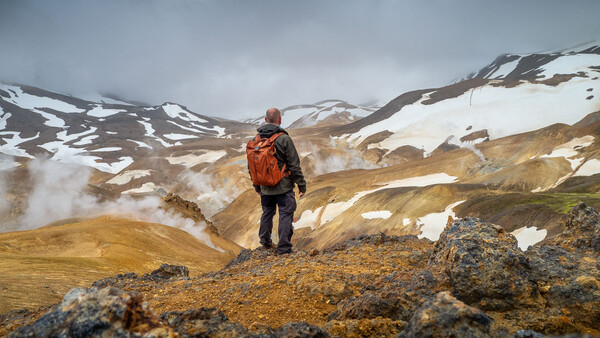 Hiker in orange backpack standing on rocky outcrop overlooking geothermal landscape with snow-capped mountains