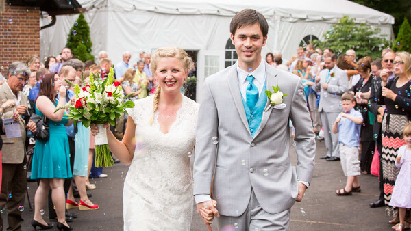 Bride and groom walk hand-in-hand through a tent, smiling at guests lining the aisle