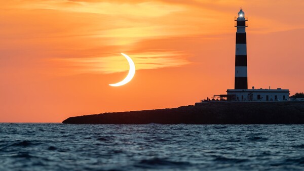 Crescent moon silhouetted against orange sunset with striped lighthouse on rocky island