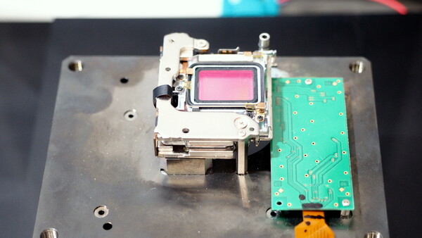 Disassembled camera internals on a metal plate: a metal-framed module with a pink-tinted sensor beside a green circuit board