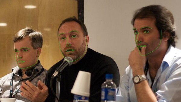 Three men on a panel, center speaker talking into a microphone, green face-tracking outlines overlaid on their faces.