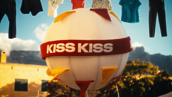 Oversized white balloon with red 'KISS KISS' band floating above landscape with clothesline of garments