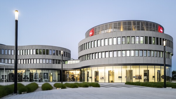 Curved modern building at dusk with red Leica logo signs on its upper level and an illuminated glass ground floor.