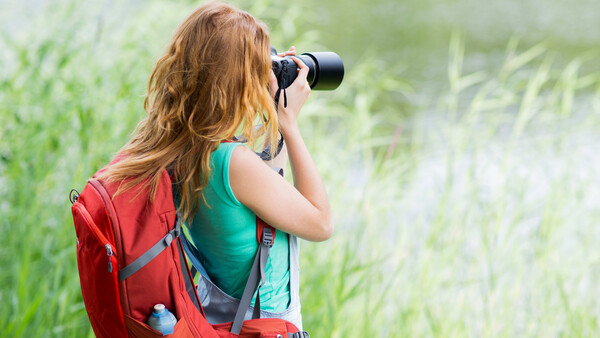 Woman with red backpack photographing outdoors with telephoto lens against blurred green field background