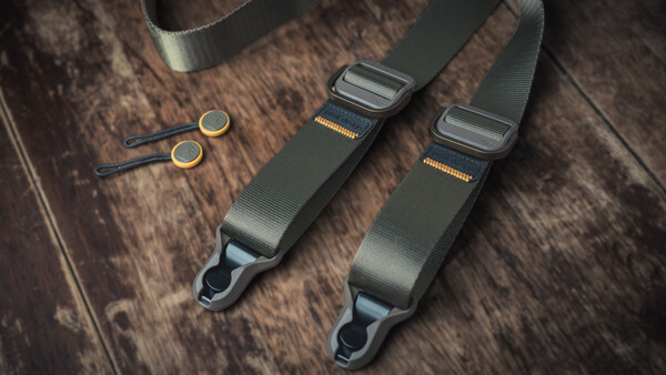 Two black camera straps with metal connectors and two small brass eyelets arranged on wooden surface
