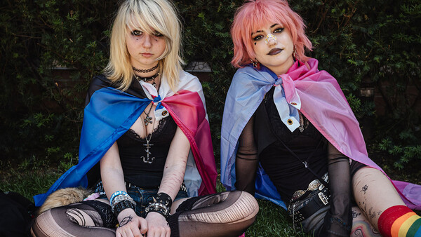 Two cosplayers in coordinated black outfits with flowing capes sitting on grass against greenery