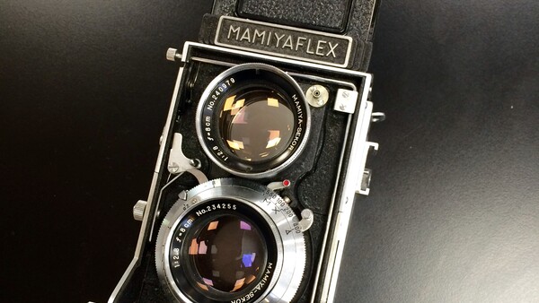 Vintage Mamiyaflex twin-lens reflex camera with a black body, chrome lens barrels, and waist-level viewfinder hood, resting on a dark surface.