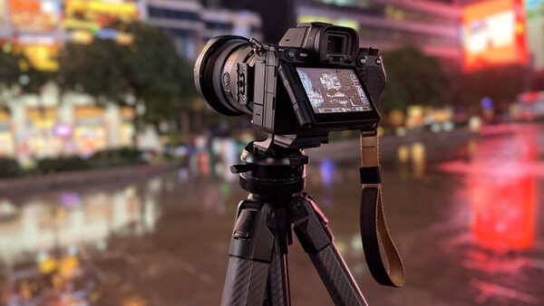 Mirrorless camera with a lens mounted on a carbon fiber tripod, rear screen angled out, set on a wet city street at night.