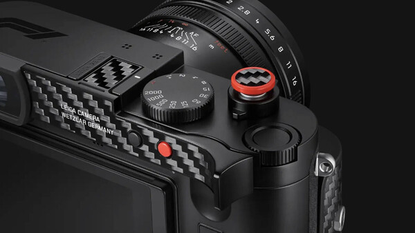 Close-up product shot of a black Leica camera's top plate and carbon-fiber grip, with a red-ringed shutter release and speed dial.