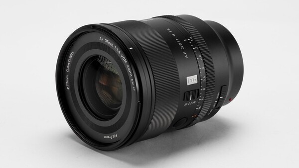 Black 35mm f/1.4 autofocus camera lens angled on a light gray studio backdrop, showing its front element and barrel markings
