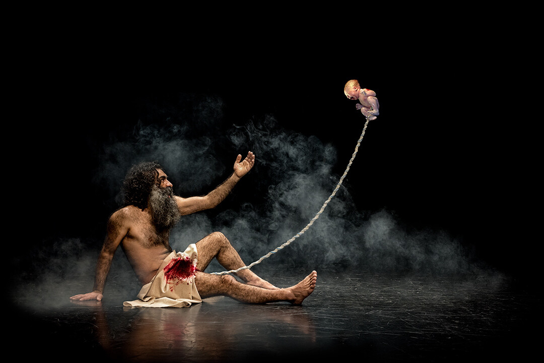 Shirtless man with long hair sitting on wet ground, arm extended upward holding a rope with a floating head