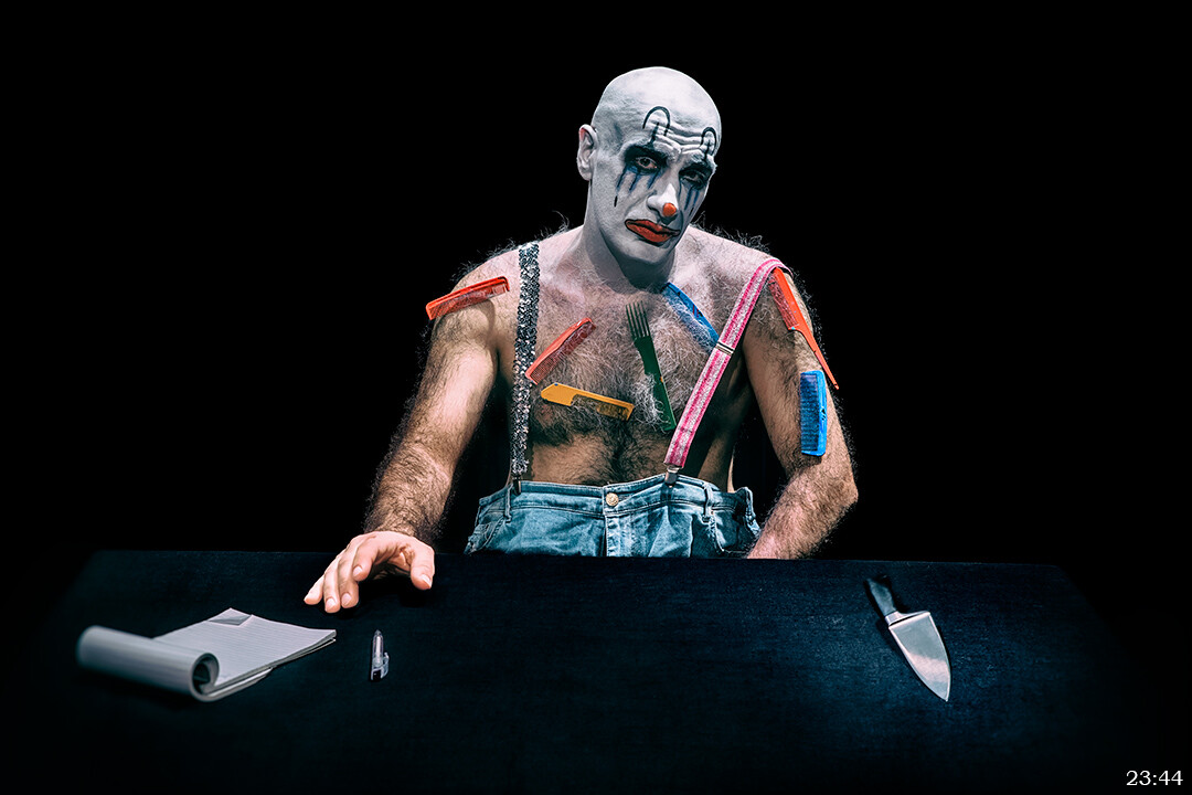 Bald man with white and blue face paint and colorful body paint sits at dark table holding papers