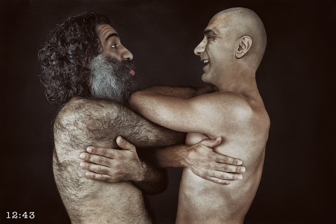 Two shirtless men with facial hair engaged in playful physical interaction against dark background