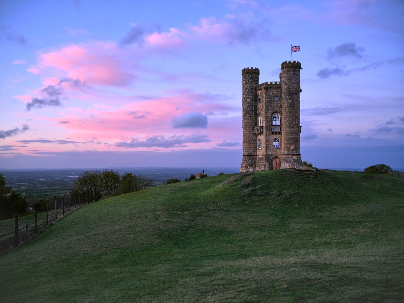 Stone tower with crenellated turrets and a Union Jack flag on a grassy hilltop at dusk under a pink and blue sky