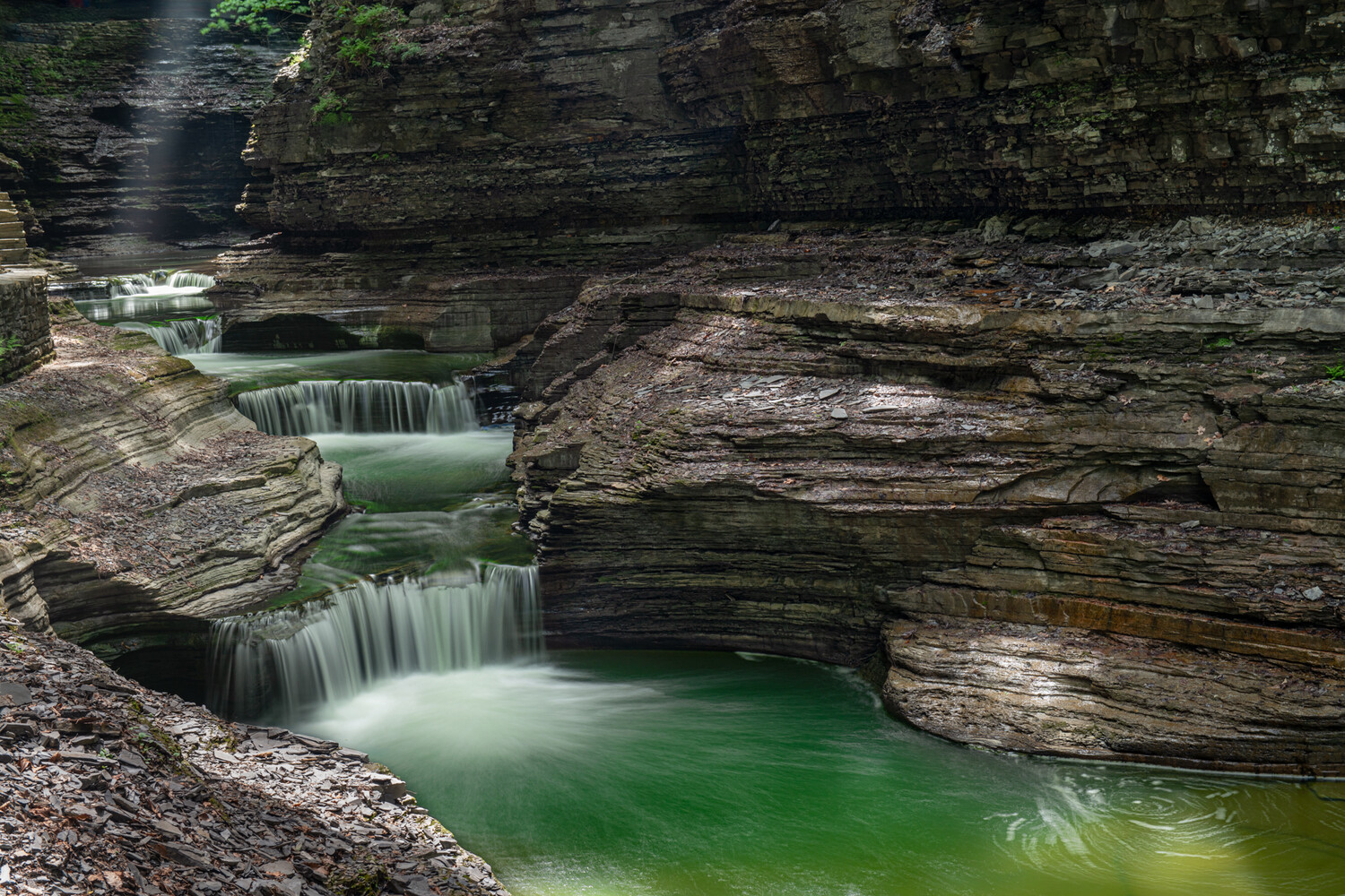 Cascading waterfalls flow into emerald-green pools carved through layered slate rock formations