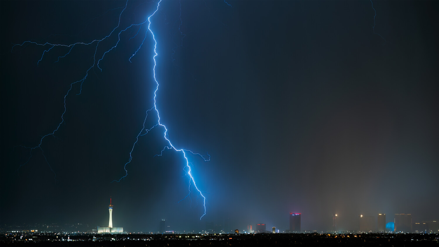 Brilliant lightning bolt striking vertically across a dark stormy sky above an illuminated city skyline