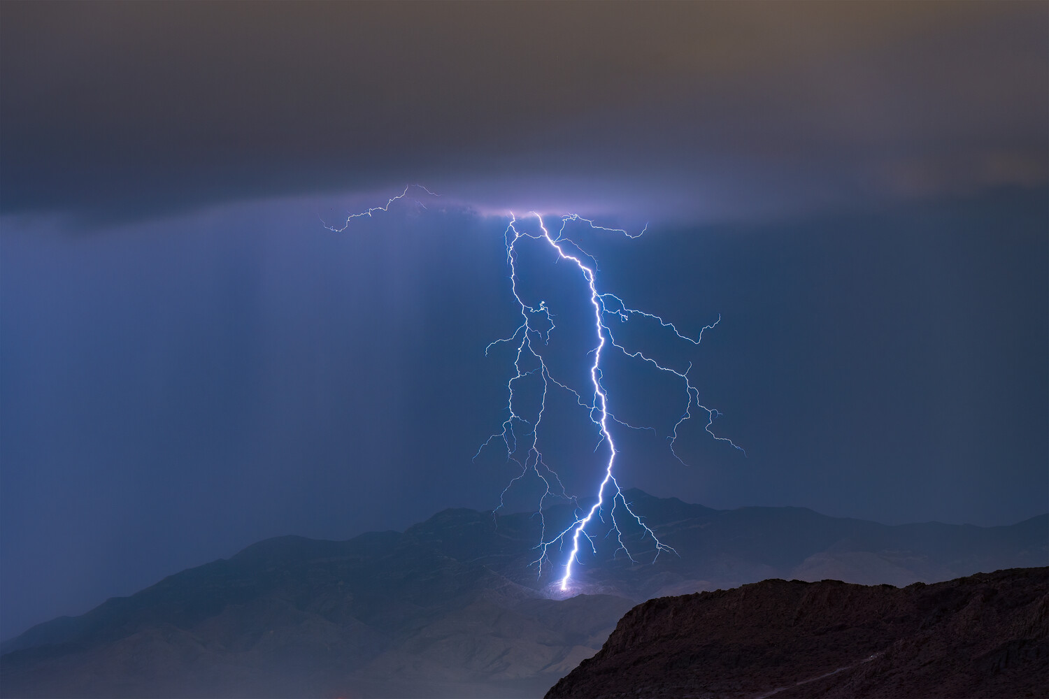 Dramatic lightning bolt strikes from storm clouds above silhouetted mountains at dusk
