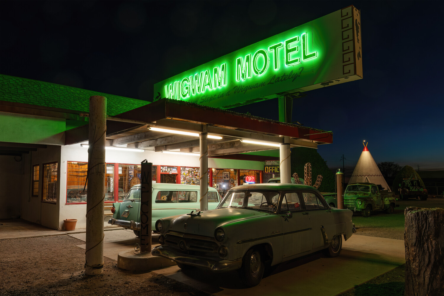 Vintage motel with neon green sign and classic 1950s car parked under a covered entrance at night