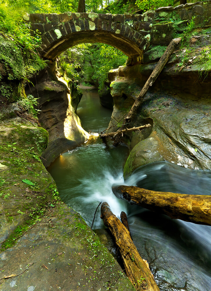 Stone arch bridge spanning a flowing stream surrounded by lush green foliage