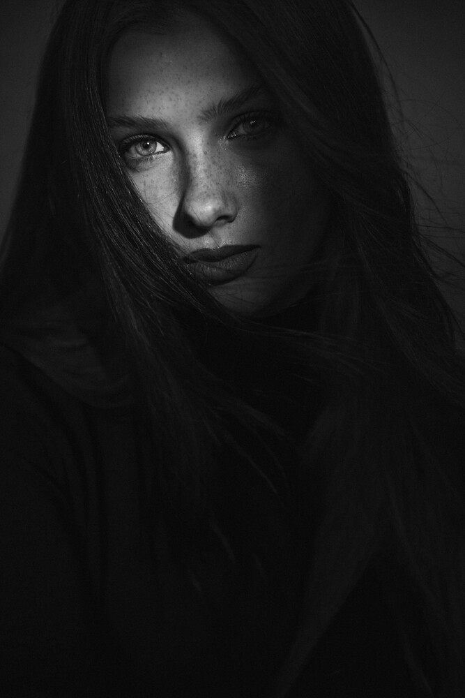 Woman with dark hair wearing a black hood, photographed in close-up against a dark background