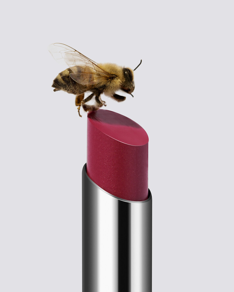 A honeybee perched on the angled tip of a deep red lipstick, set against a plain light gray background.