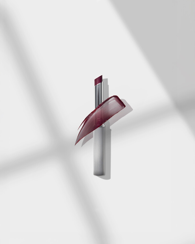 Burgundy lipstick with a glossy plum swatch sweeping off the tip, set against a white backdrop with diagonal shadows.