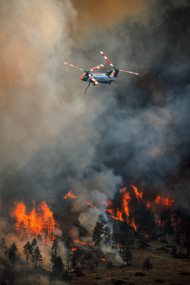 Helicopter conducting aerial firefighting operations above active wildfire with flames and smoke