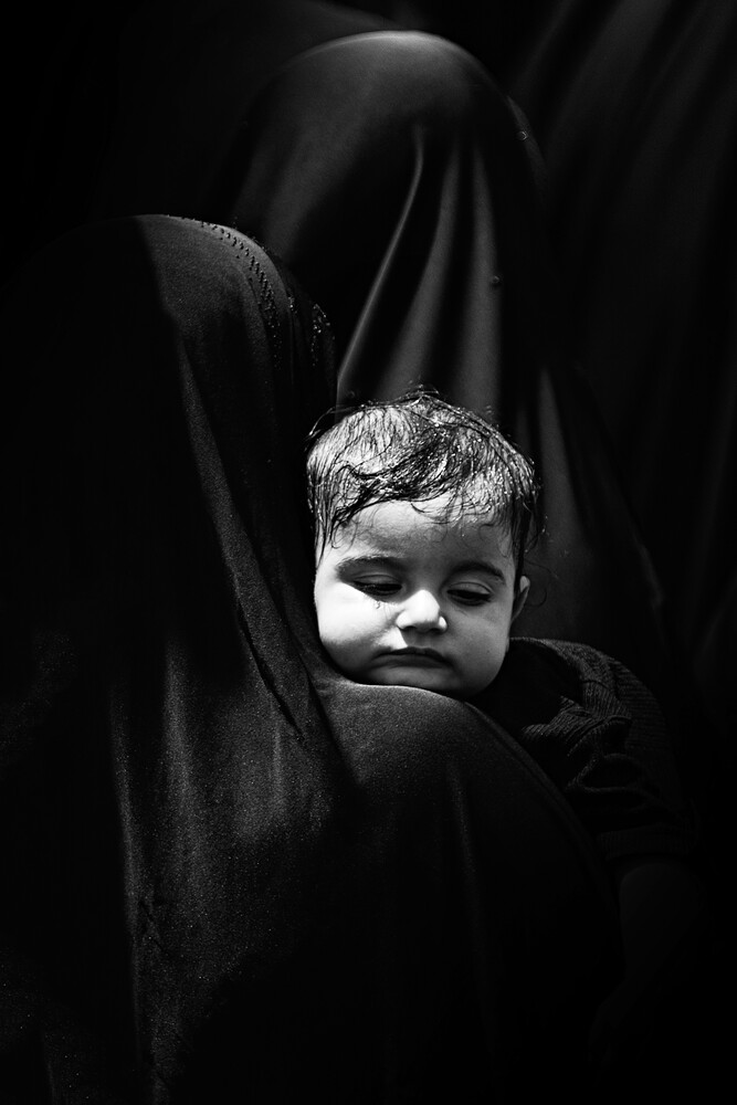 Young child's face emerging from dark draped fabric, high-contrast black and white portrait