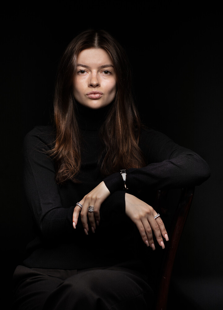 Woman with long dark hair in black clothing against dark background, seated with hand positioned on knee
