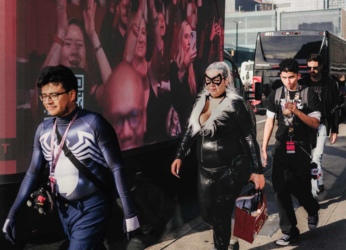 Cosplayers in Venom and Black Cat costumes walking past a large outdoor screen showing a cheering crowd, with a bus behind them.
