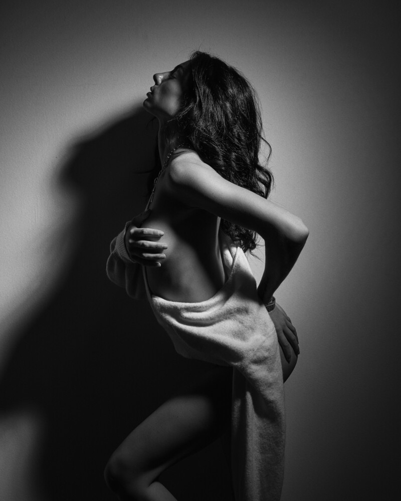 Shirtless man with long dark hair hugging himself against a gray wall in dramatic black and white studio lighting