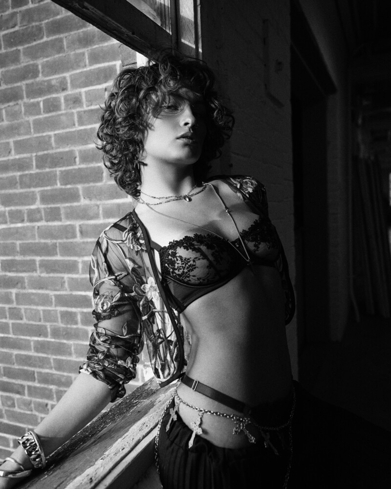 Woman with curly hair in patterned shirt and bikini top posing against brick wall in black and white