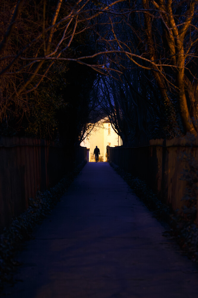 Narrow paved path at night with bare trees overhead, leading toward a glowing archway where a lone figure stands in silhouette.