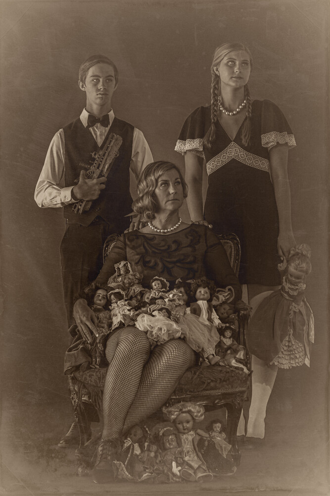 Sepia-toned staged group portrait of three people and a doll, with a seated woman surrounded by vintage dolls on an ornate chair.